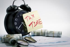 Alarm clock with sticky note reading Tax Time beside money rolls and financial papers, symbolizing IRS tax problems and filing deadlines.