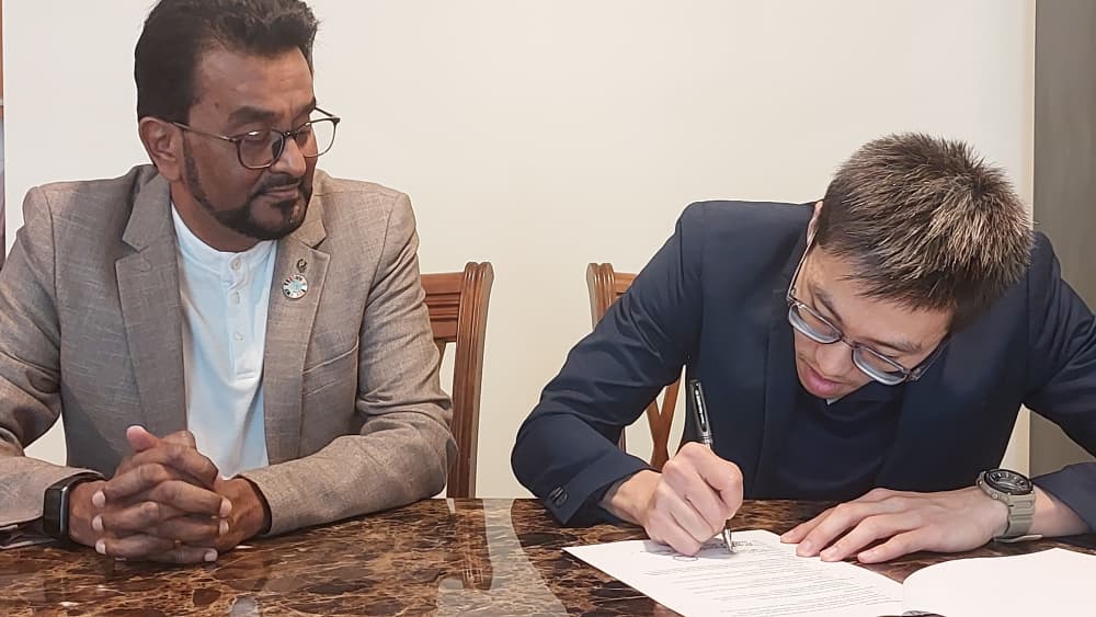 Two representatives at a signing table, one witnessing while the other signs the memorandum of understanding.