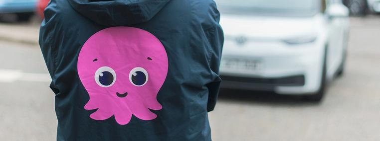 Person seen from the back wearing a dark blue jacket with a pink Constantine the Octopus printed on it, in the further background a white electric vehicle