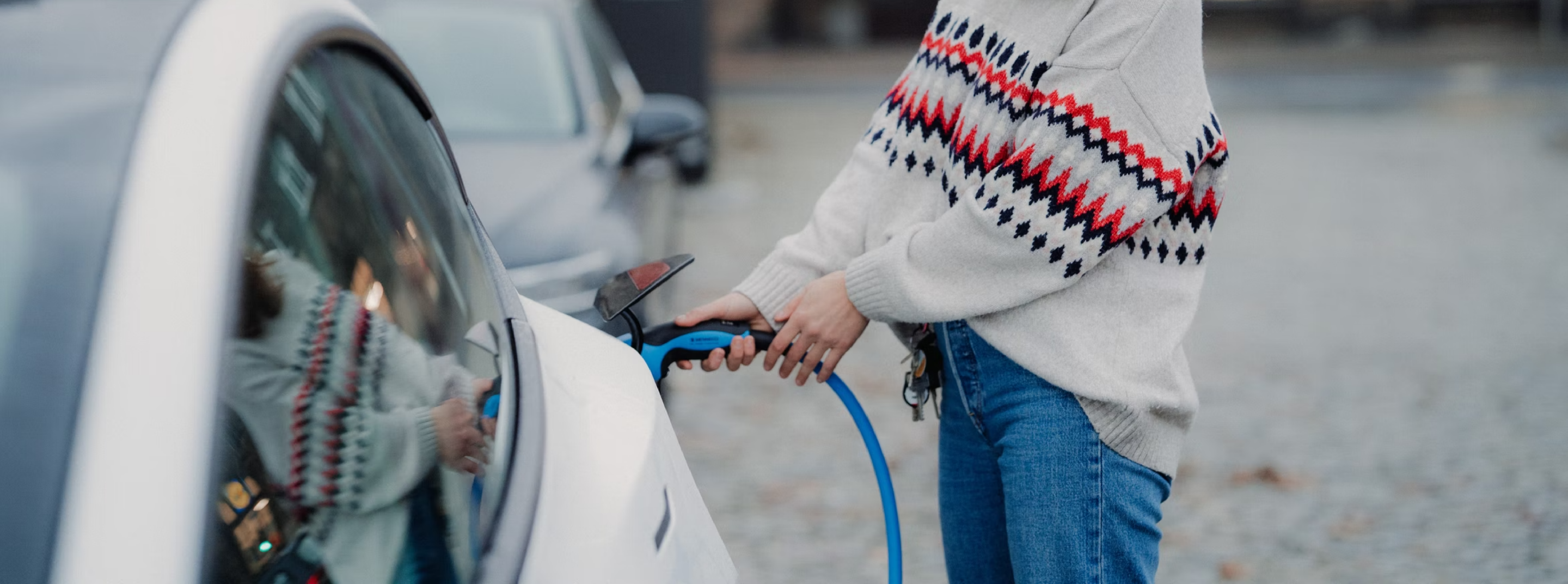 Person in white knitted jumper charging a white EV with a blue charging cable