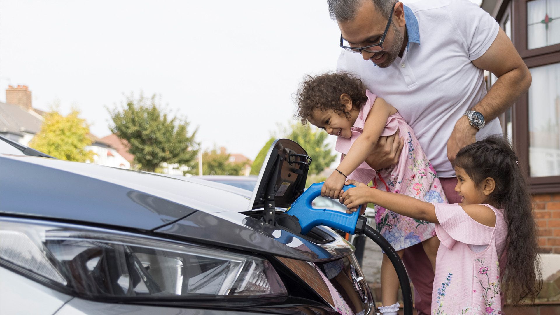 A person is holding a small child up to an electric vehicle to help them charge and a second child is standing on the ground holding on to the electric vehicle charger as well