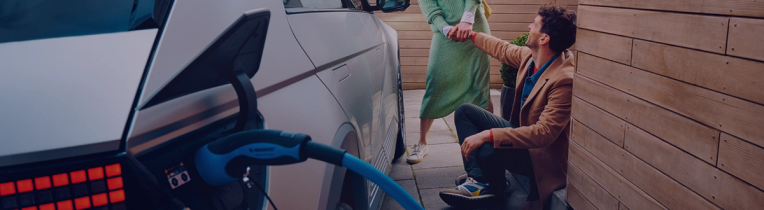 A grey electric vehicle is charging next to a house where a man is sitting down and reaching up to a person wearing a dress by the hand