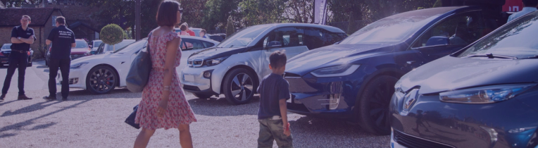 Person looking at their phone and in the background is a light grey electric vehicle charging by an EV charger