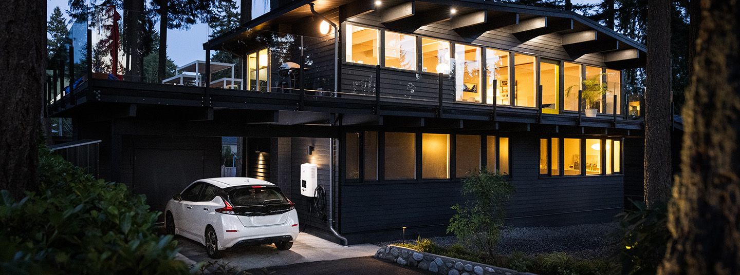 Nissan Leaf in white parked out side a modern house next to a electric vehicle charger with lights on in the kitchen