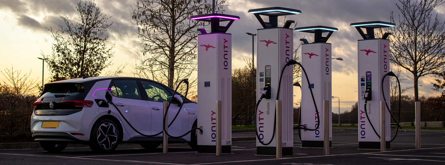 white electric vehicle charging next to several white vehicle charging points with a sunset in the background