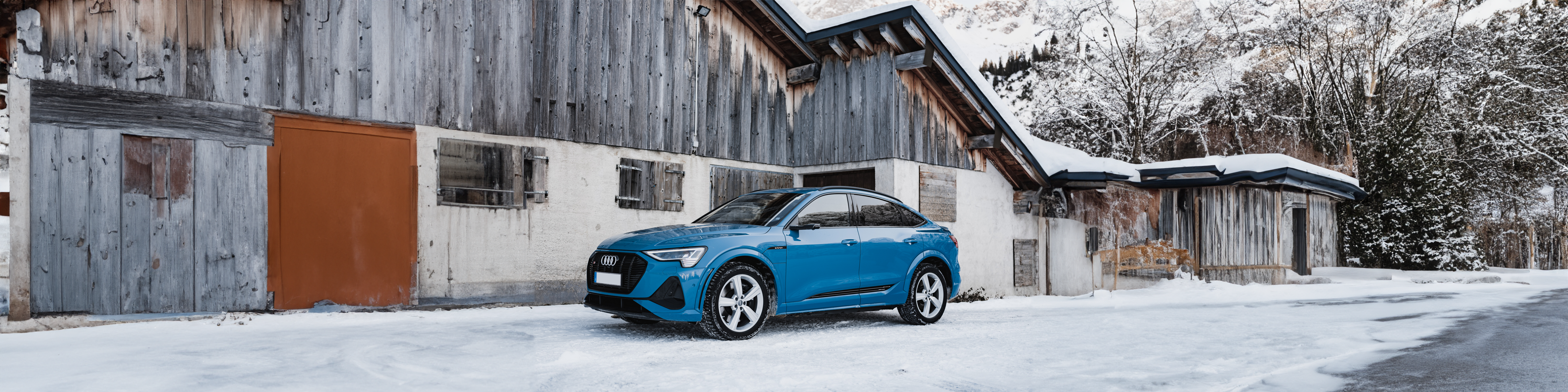 A blue Audi E-tron S Sportback parked in the snow in front of some buildings
