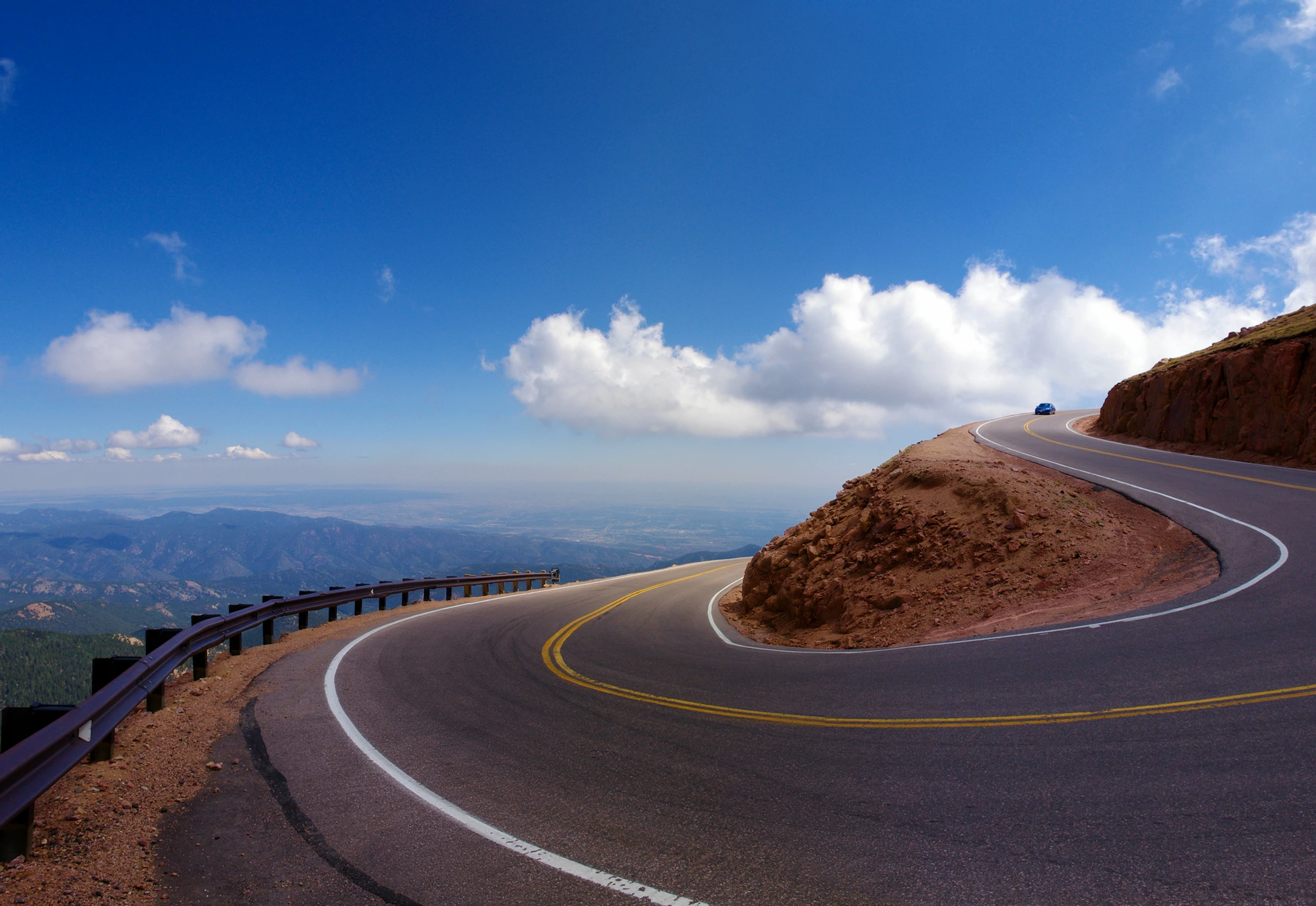 Image of a road bending around a curve with a blue sky and white clouds on a high altitude with views over mountains and hills
