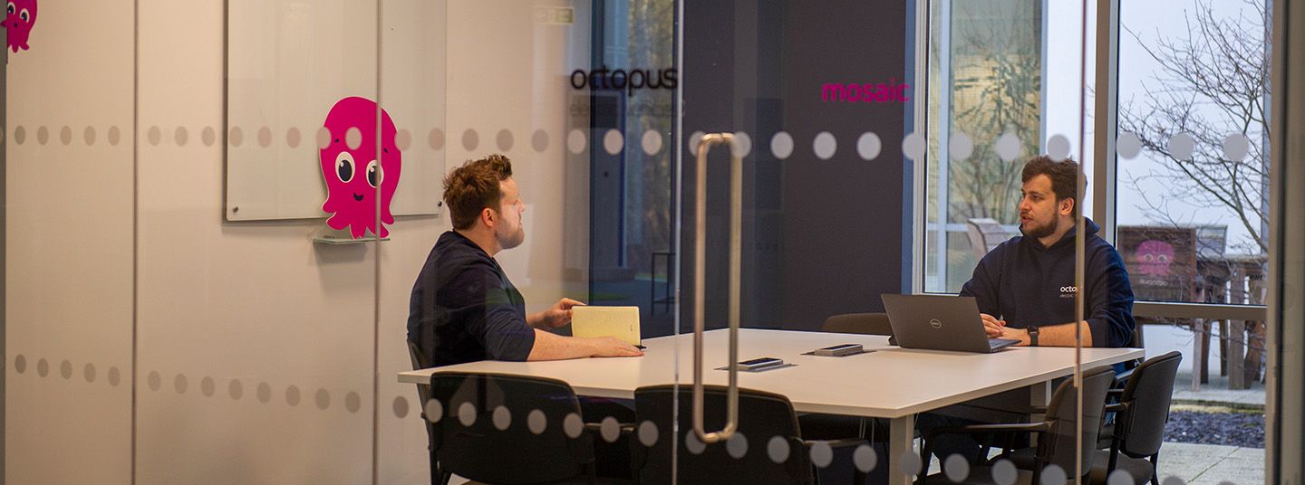 Two people sitting in a meeting room seen through a glass door with a pink Octopus on the whiteboard