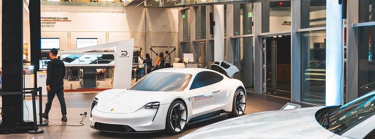 a white electric vehicle with the make of Porsche stands in an exhibition hall with a person dressed in black walking away from it