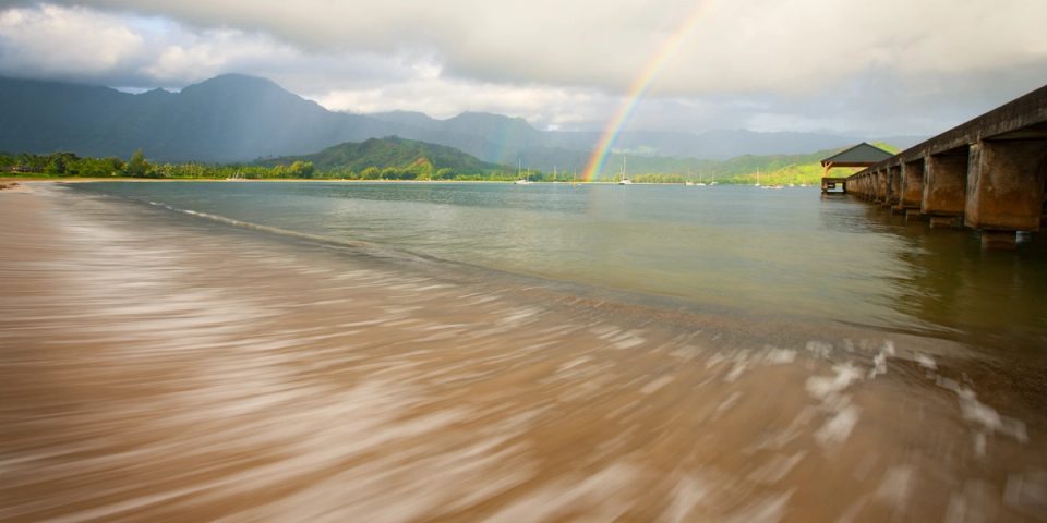 Hanalei Bay on Kauai under broken cloud, a rainbow over the green ridges and the pier at right, calm water and a sandy beach in the foreground.