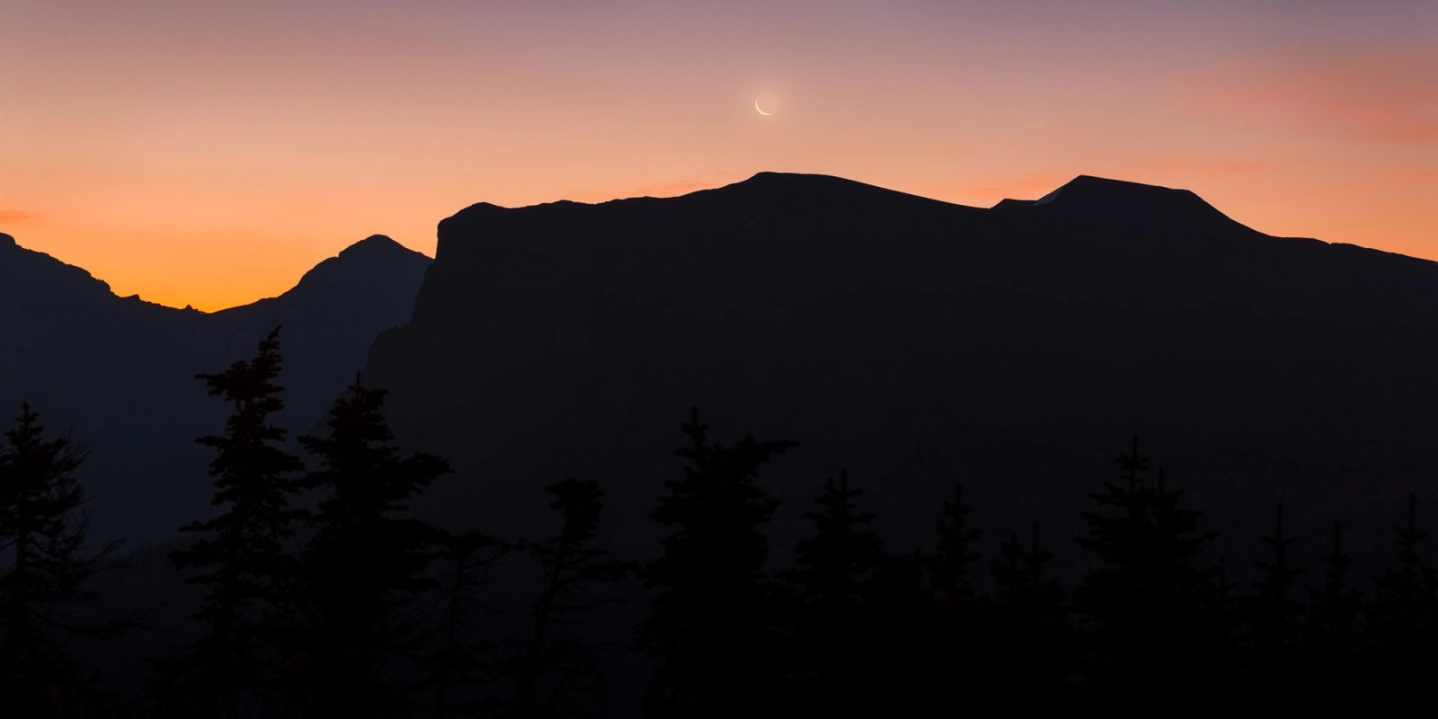 A thin crescent moon above layered, silhouetted mountain ridges at dawn, with an orange glow along the horizon and dark spruce tops in the foreground.