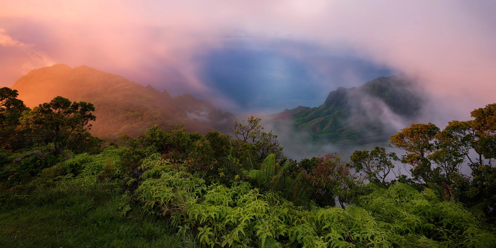 Sunset light glowing on low clouds blowing through Kalalau Valley, with the green ridges of the Na Pali coast and a view down to the ocean.