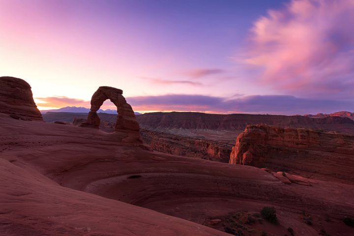Delicate Arch in shadow against a pink and purple sunrise sky, with the La Sal Mountains on the horizon and a sweeping bowl of red slickrock in the foreground.