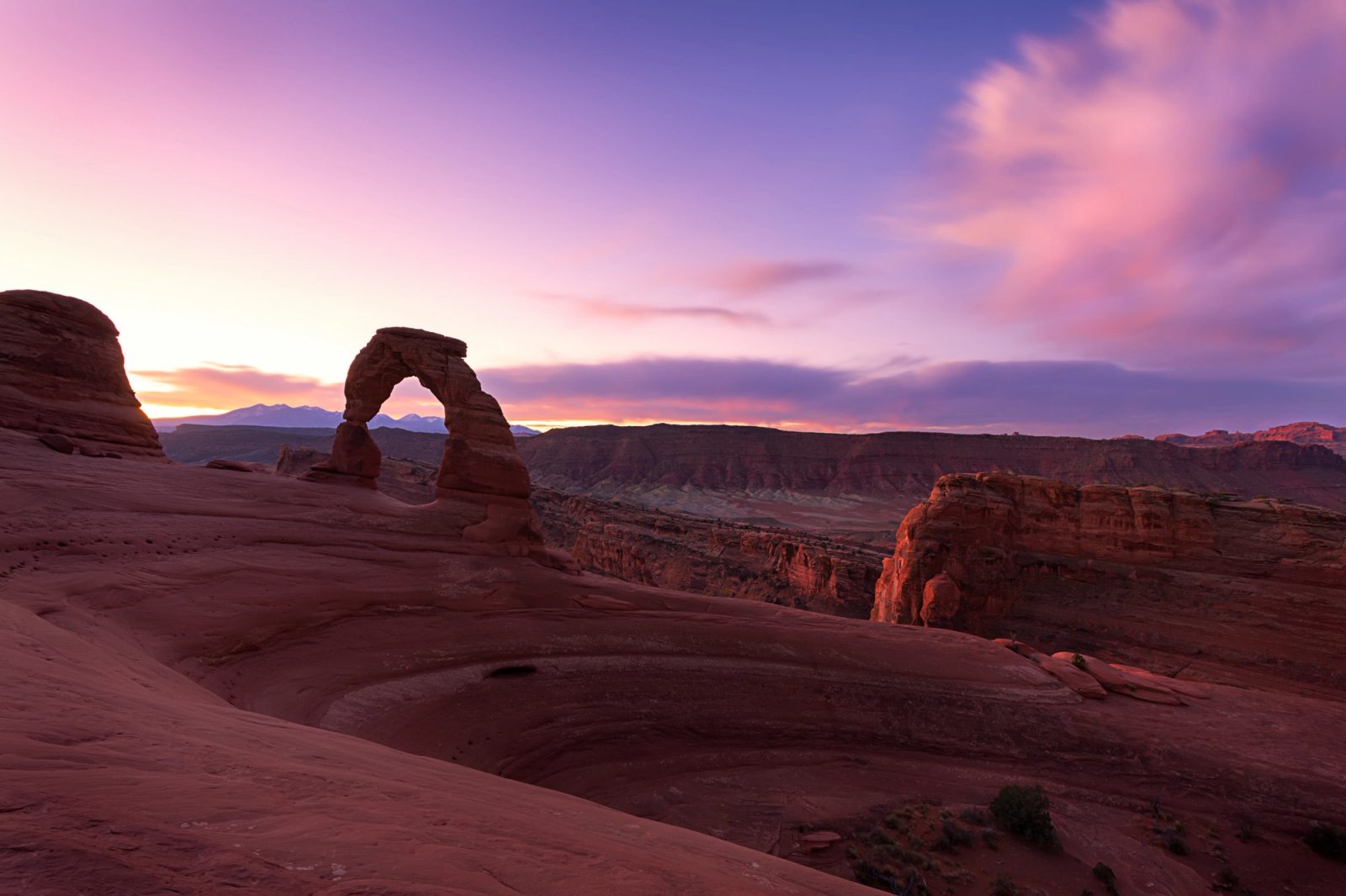 Delicate Arch in shadow against a pink and purple sunrise sky, with the La Sal Mountains on the horizon and a sweeping bowl of red slickrock in the foreground.