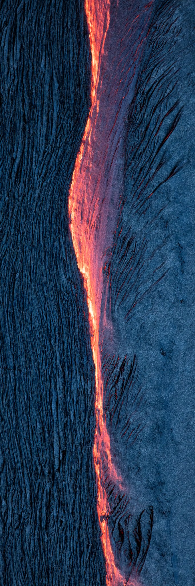 An aerial view of a glowing orange seam of molten lava running between two sheets of crusted pahoehoe, one smooth and silvery, the other ropy and dark, the cooled crust a dark blue-gray.