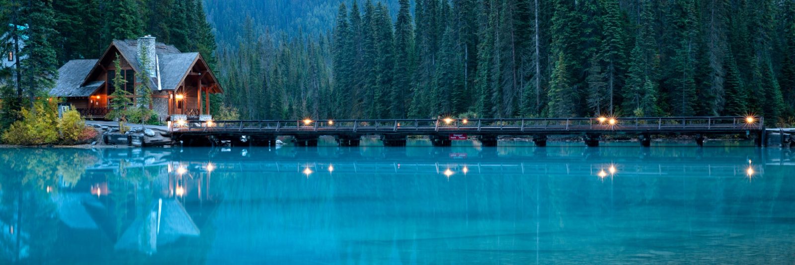 Emerald Lake Lodge and footbridge at blue hour, warm lights reflecting in still turquoise water under a forested shore, Yoho National Park.