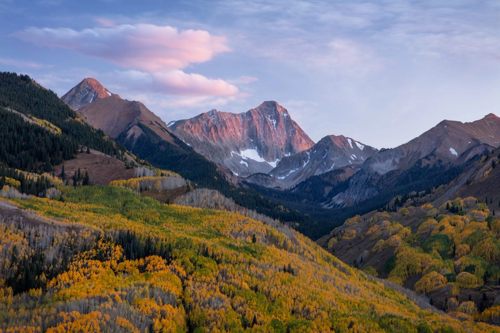 Capitol Peak lit pink at sunset above a dark timbered valley, with a cloud overhead catching the same light and hillsides of yellow and green aspen in the foreground, Colorado.
