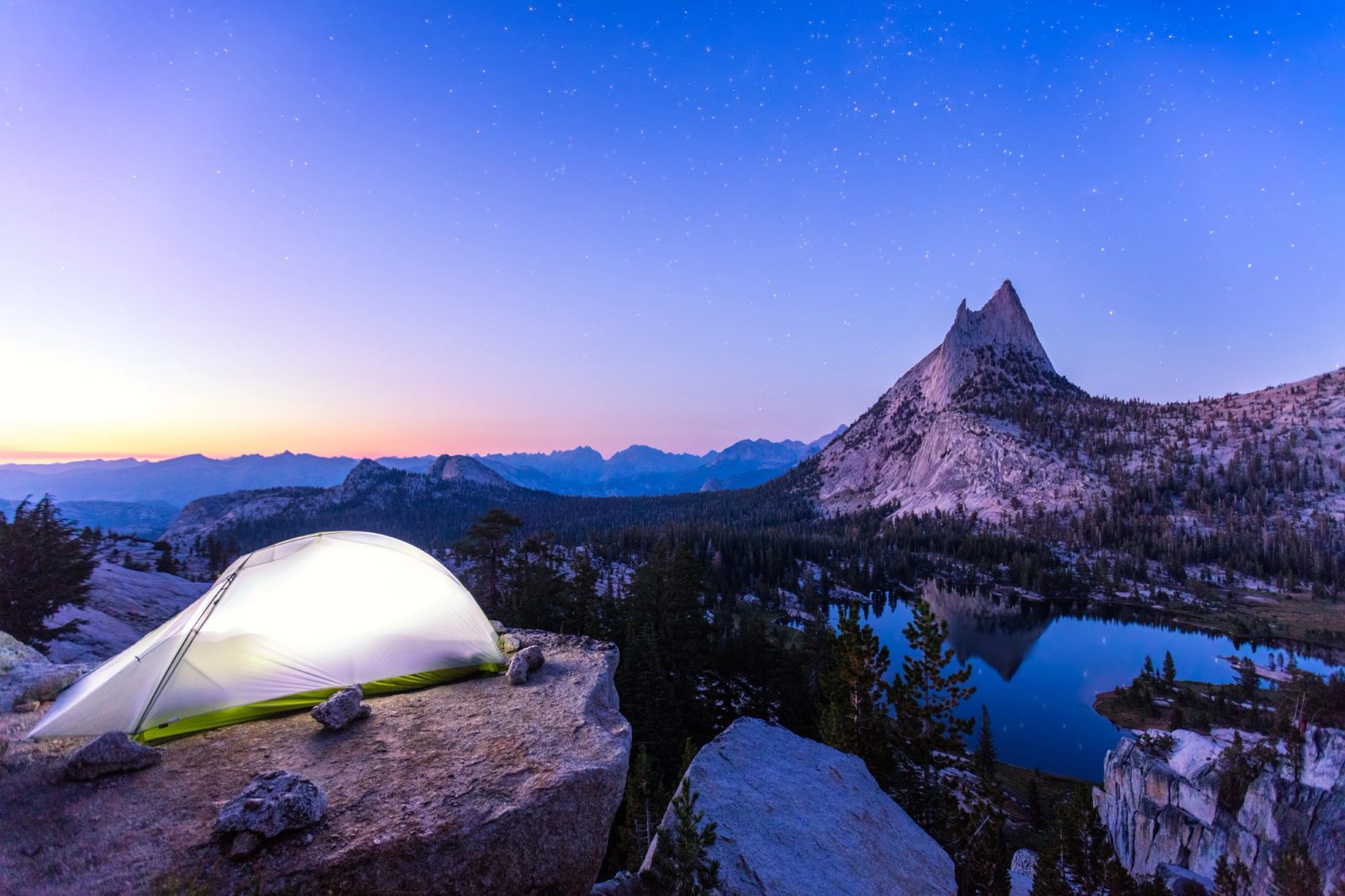 A lit tent on granite above Upper Cathedral Lake, Cathedral Peak reflected in the water under a starry sky.