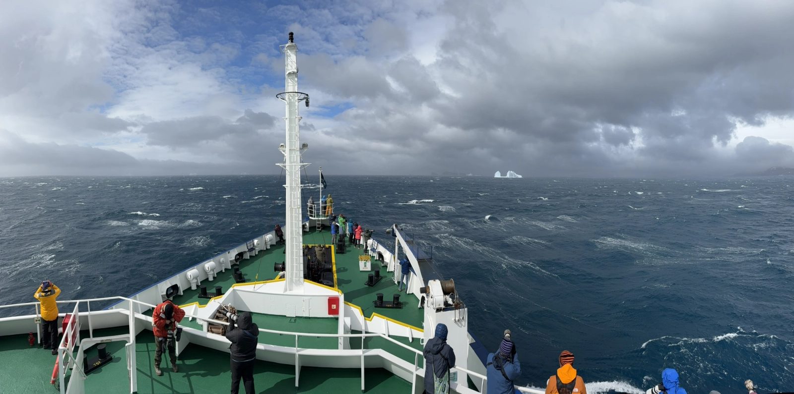 Panorama from the ship's upper deck of passengers in bright jackets lining the bow, watching a distant iceberg on a rough, dark blue sea under heavy clouds.