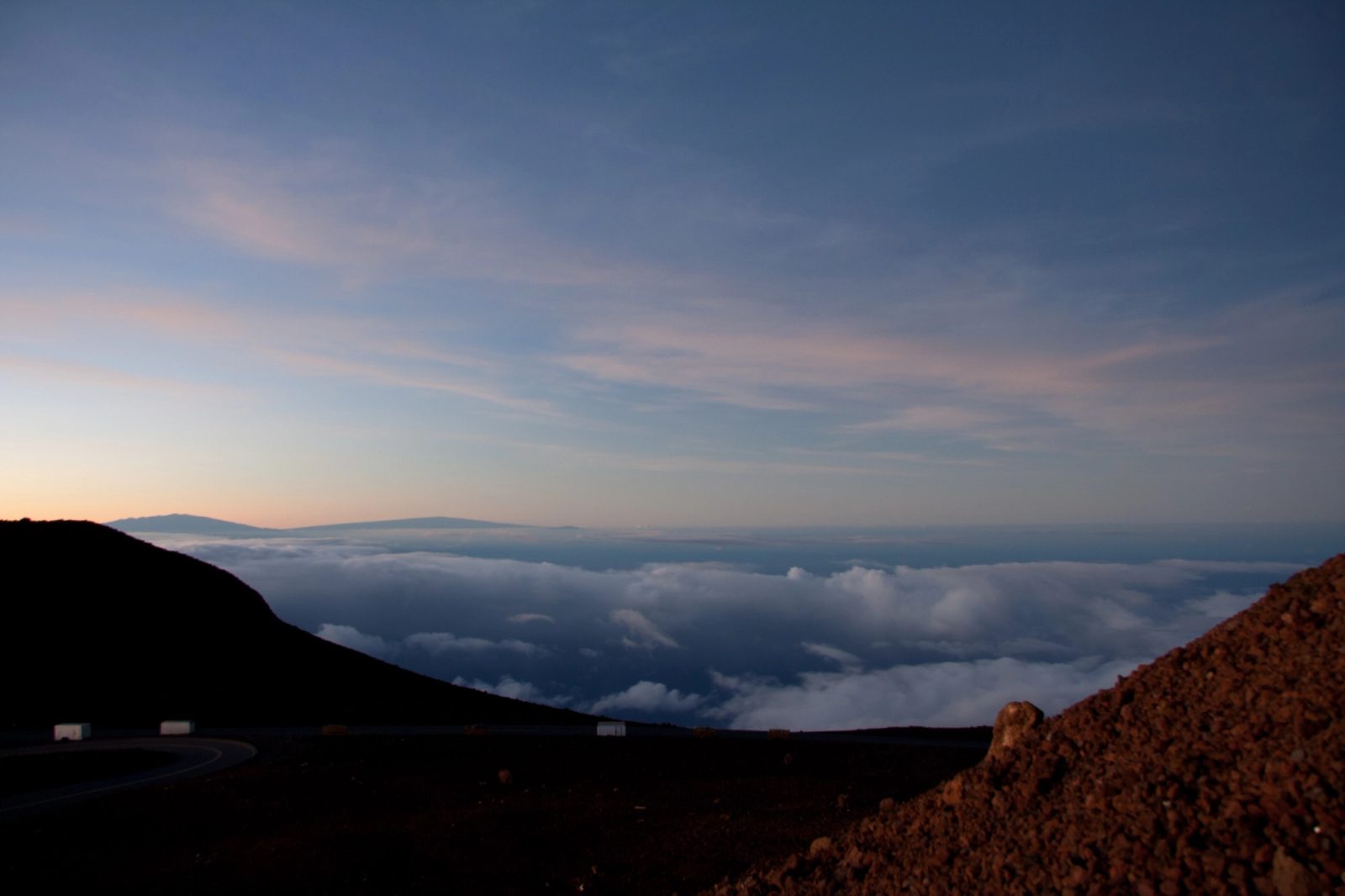 Dusk above a cloud layer from a volcano summit road, two distant peaks breaking the horizon, red cinder slope in the foreground.