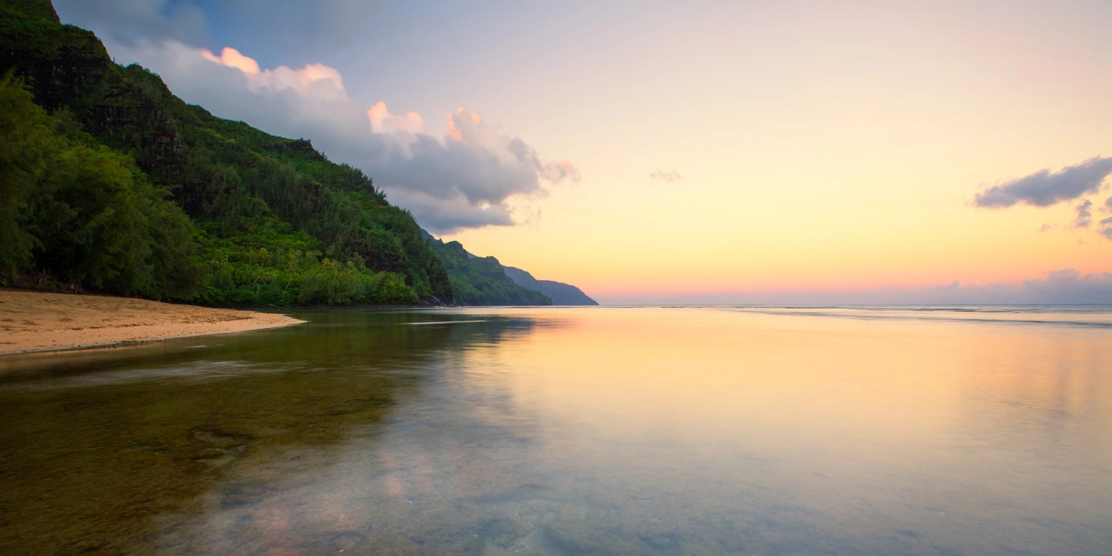 Ke'e Beach on Kauai at sunrise, with the green Na Pali cliffs on the left, sunlit clouds along the horizon, and calm, clear shallow water reflecting the warm sky.