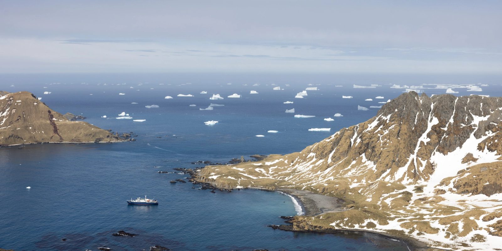 High view over Cooper Bay, with a small ship anchored in deep blue water, Cooper Island offshore, a snow-streaked ridge and dark beach in the foreground, and dozens of icebergs out to the horizon.