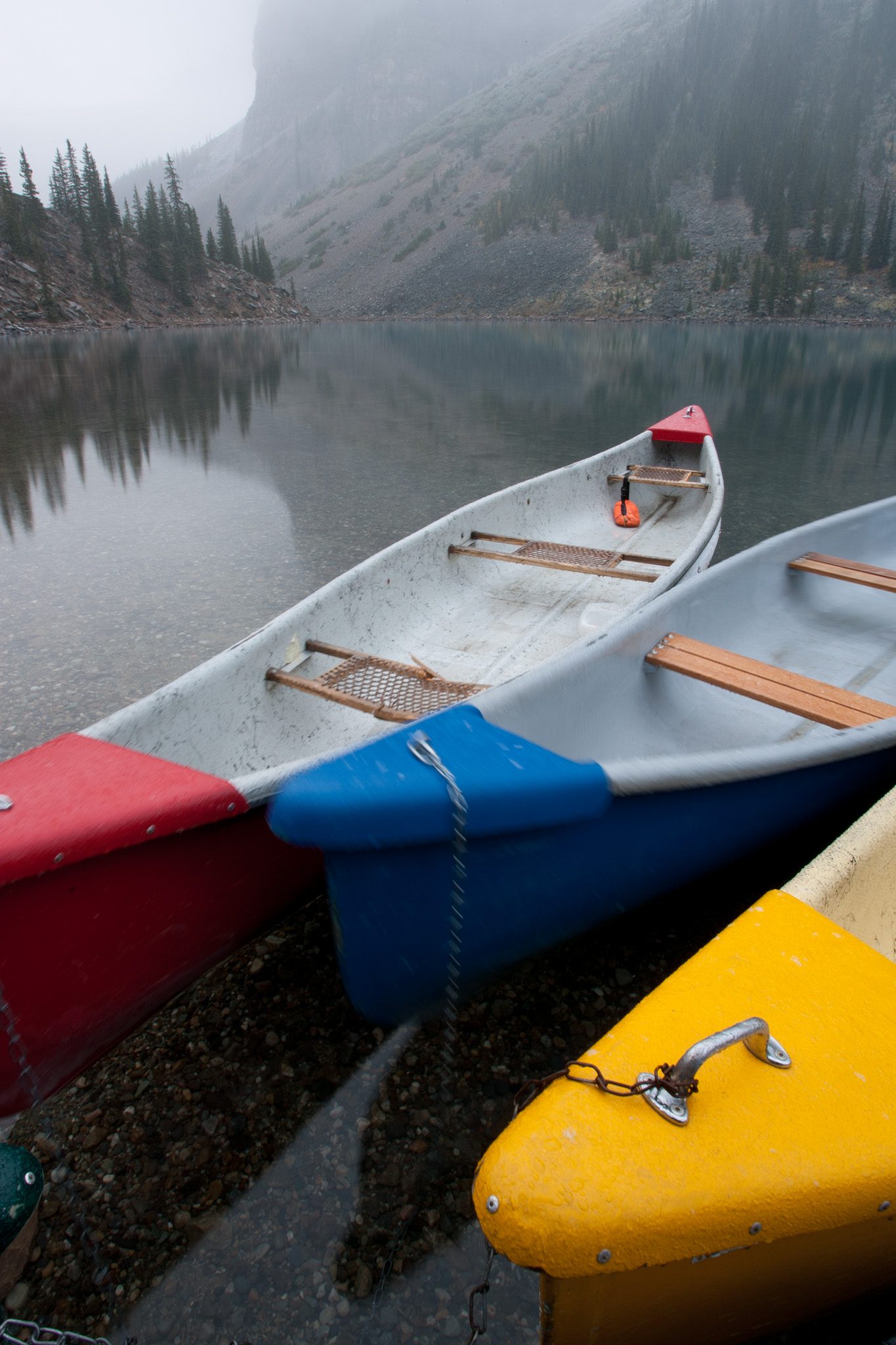 Red, blue, and yellow canoes at the edge of Moraine Lake in fog, slightly out of focus.