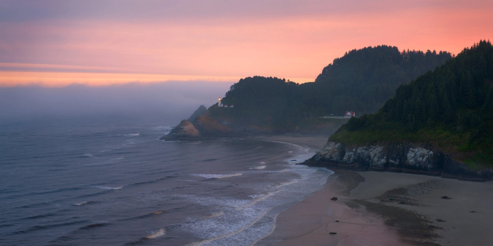 Heceta Head Lighthouse shining from a forested headland above the Pacific, with fog moving in under a pink sunrise sky and a curving beach and rocky point in the foreground.