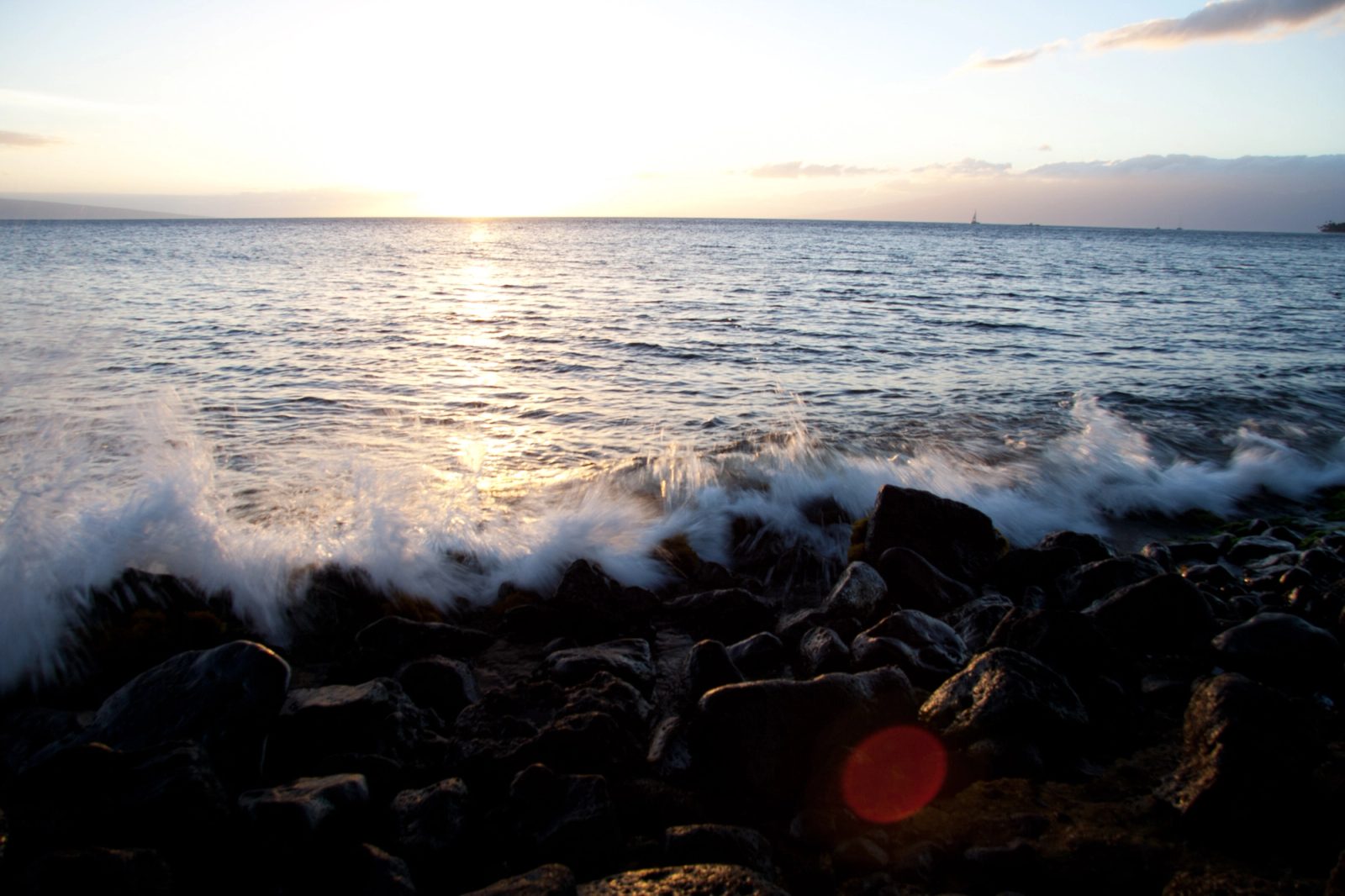 Sunset over the ocean from a rocky lava shoreline, a wave breaking white across the rocks, a sailboat small on the horizon.