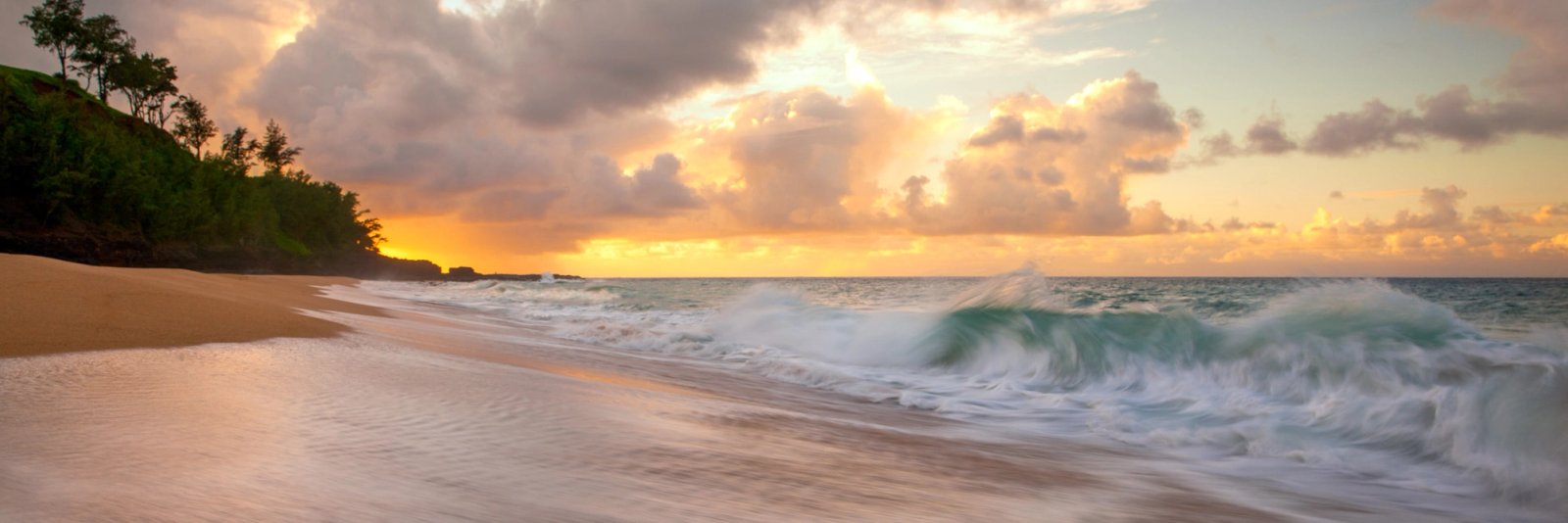 A wave curling over in green water at Secret Beach, Kauai, with foam washing up the sand toward a tree-covered point and warm sunset light breaking through heavy clouds.