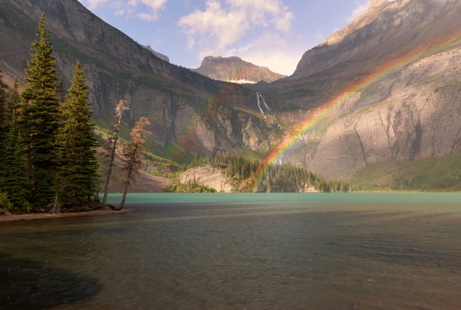 A rainbow over the headwall above Grinnell Lake, turquoise water in the foreground and spruce along the near shore.