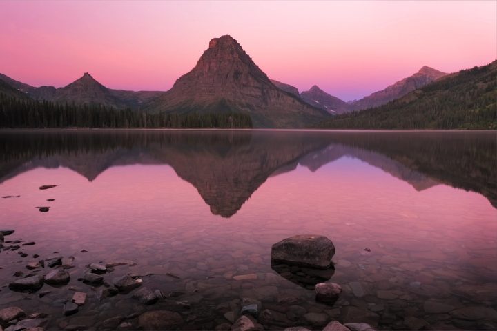 Sunrise light on Sinopah Mountain reflected in the still water of Two Medicine Lake, Glacier National Park, Montana, under a pink sky.