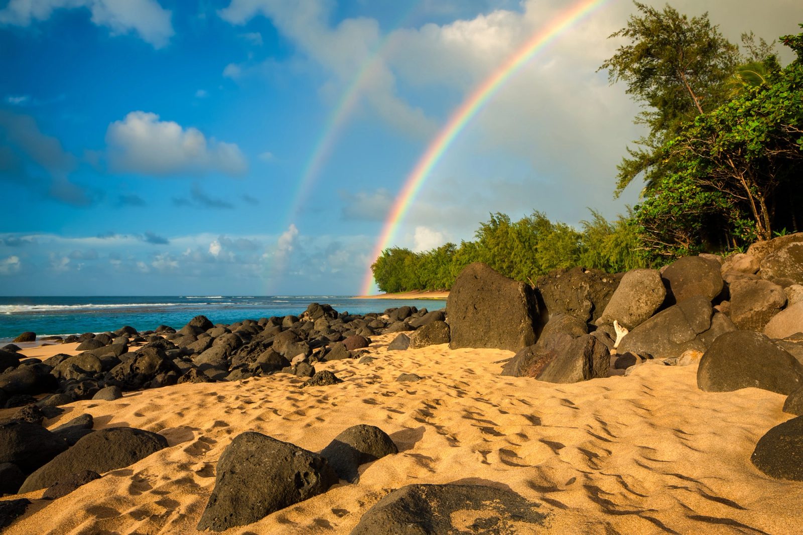 A double rainbow arcing over green ironwood trees at Ke'e Beach, Kauai, with black lava boulders on golden sand lit by low evening sun.
