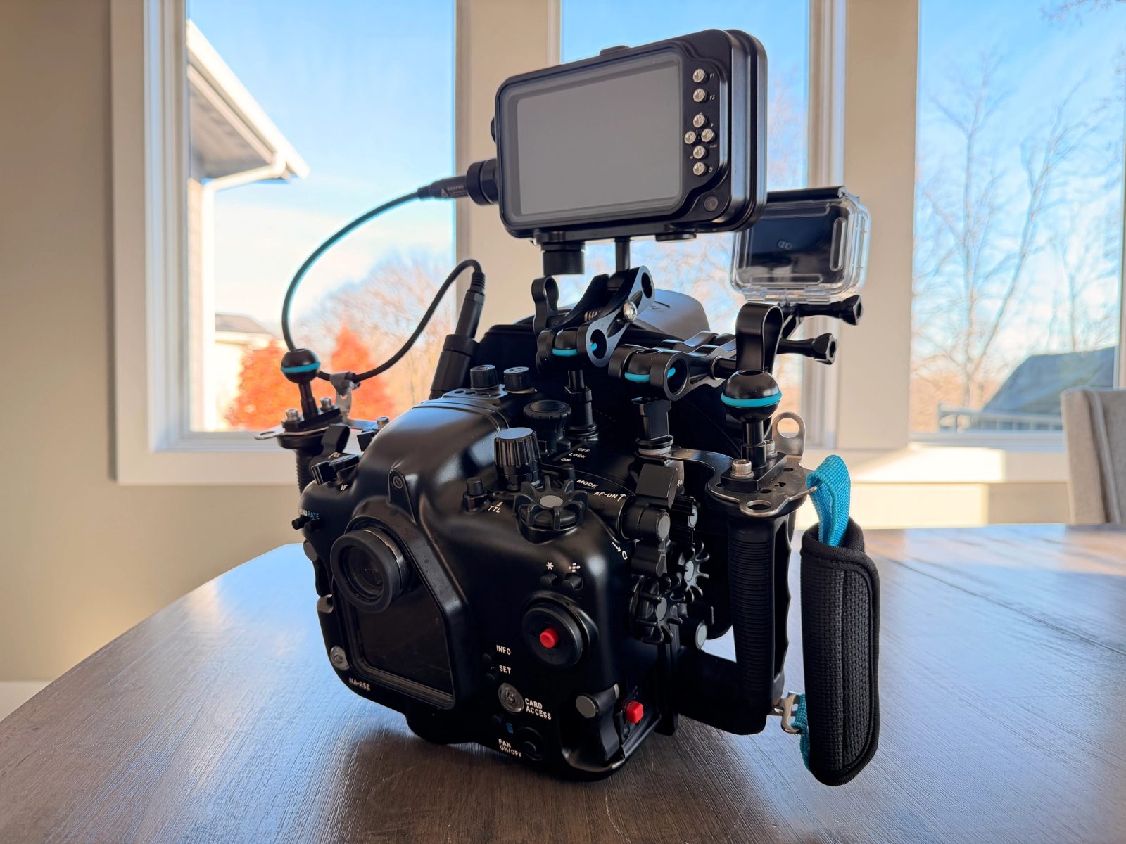 Rear of an underwater camera housing with handles, a monitor, and a GoPro, on a table indoors.