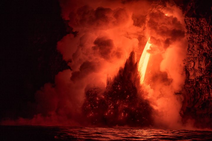 A stream of molten lava pouring from a sea cliff into the ocean before dawn, throwing up a dark burst of debris inside clouds of red-lit steam.