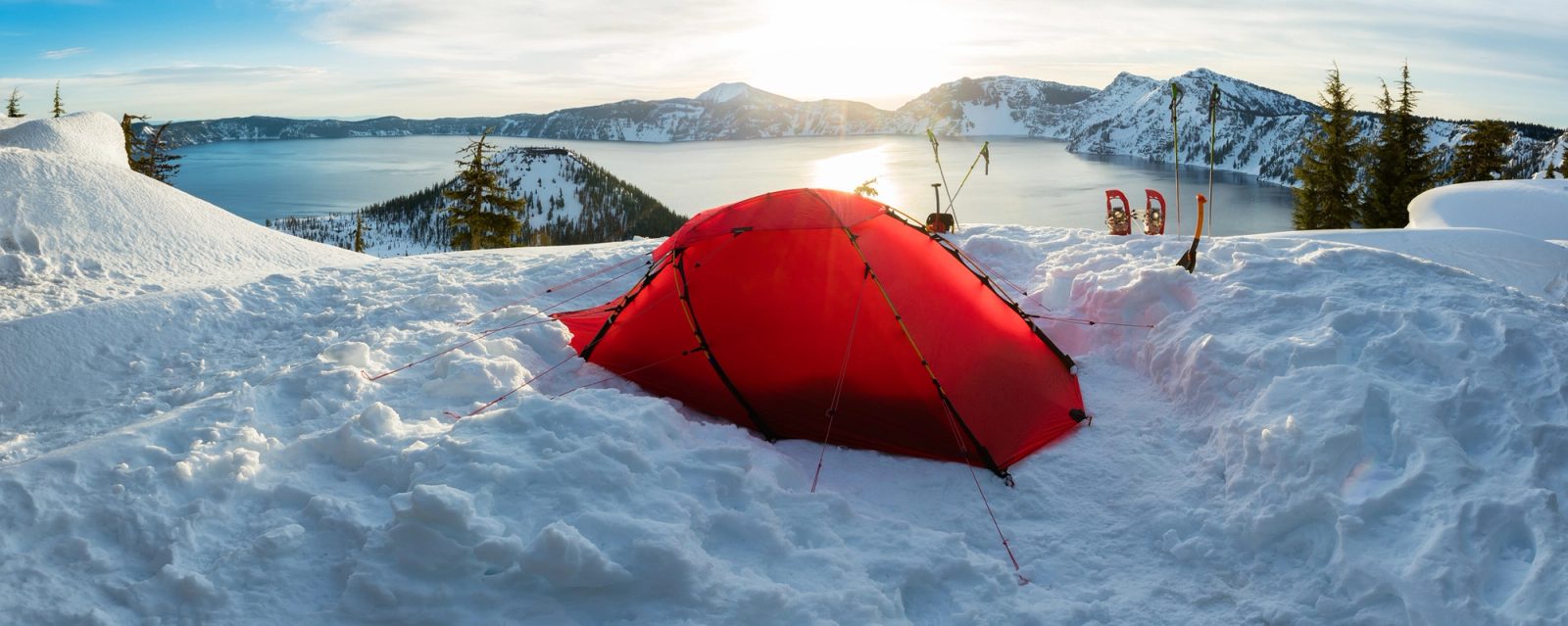 A red tent dug into deep snow on the rim of Crater Lake, Oregon, with Wizard Island and the caldera behind in evening light.