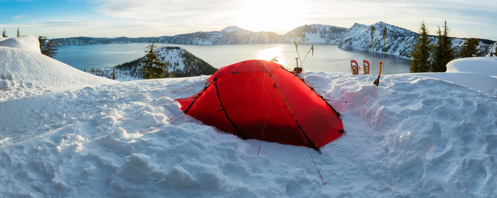 A red tent dug into deep snow on the rim of Crater Lake, Oregon, with Wizard Island and the caldera behind in evening light.