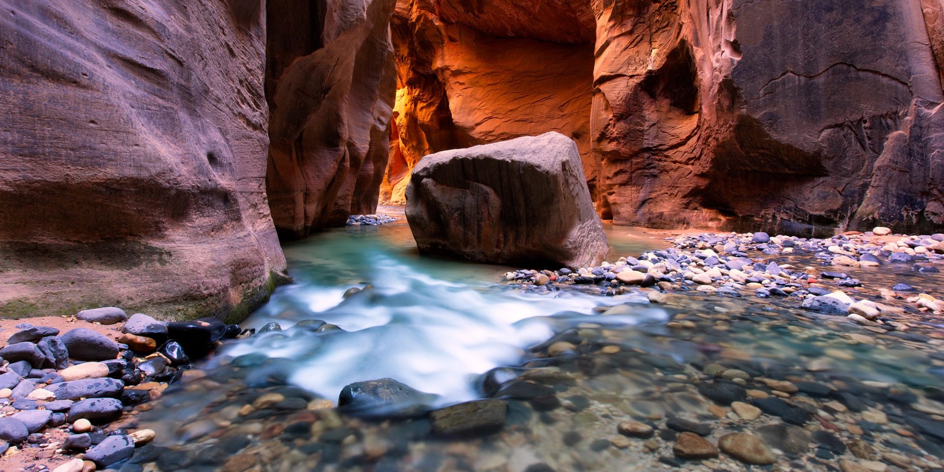Sunlit orange walls of a narrow slot canyon above turquoise water, a large boulder standing in the middle of the river channel.