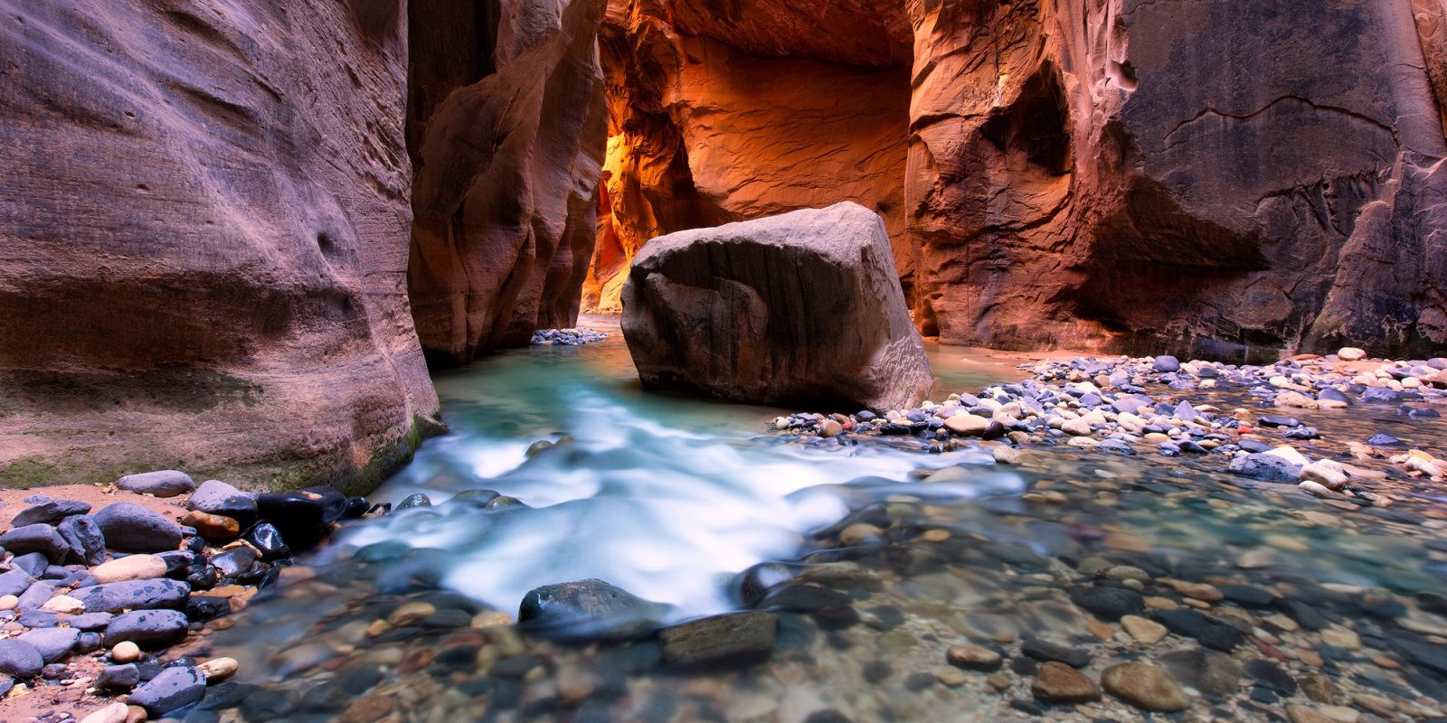 Sunlit orange walls of a narrow slot canyon above turquoise water, a large boulder standing in the middle of the river channel.