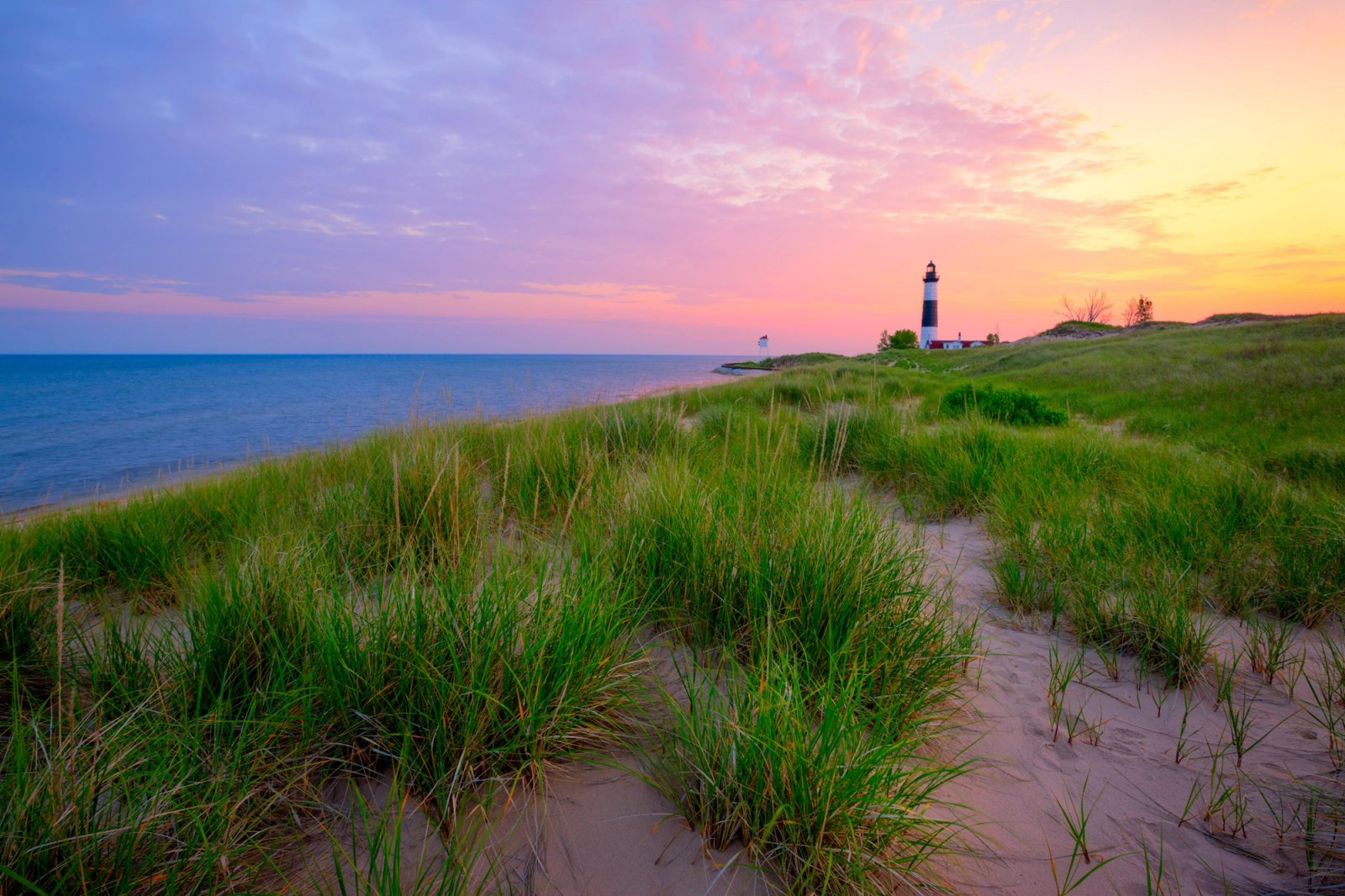 Big Sable Point Lighthouse beyond dune grass at sunrise, pink and orange sky over Lake Michigan.