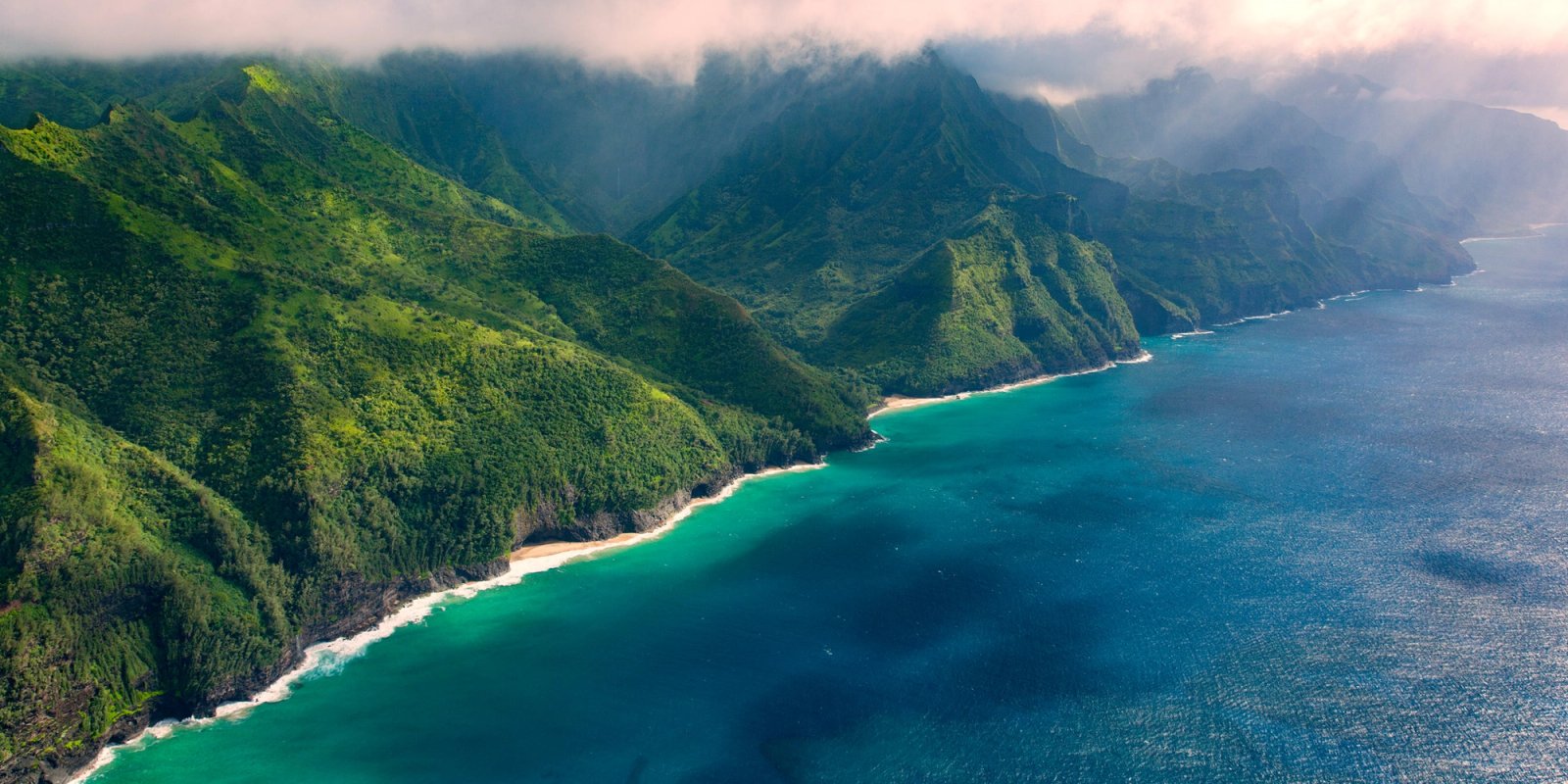 An aerial view down the Na Pali coast on Kauai with green fluted ridges, a curved beach and reef, and sunbeams falling through cloud onto turquoise water.
