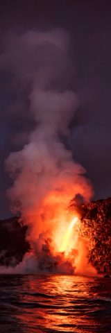 Lava firehose pouring into the ocean at Kalapana, steam plume lit orange above dark water, Big Island, Hawaii.
