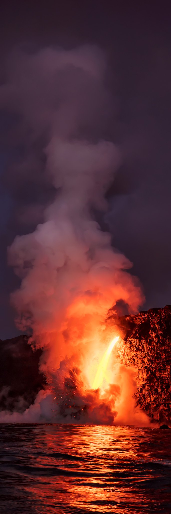 Lava firehose pouring into the ocean at Kalapana, steam plume lit orange above dark water, Big Island, Hawaii.