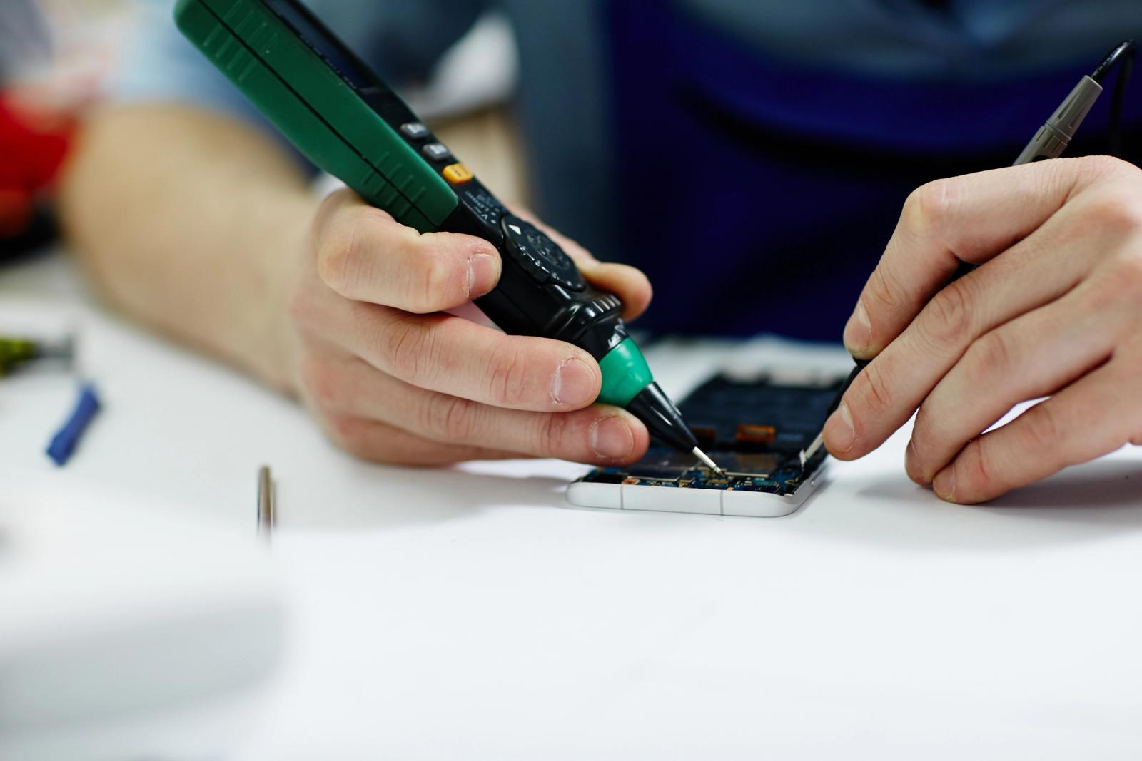 A smartphone or electronic device lays on a workbench while someone attempts to repair the internal components