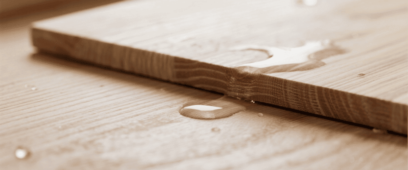 Wooden plank with water droplets on a light wood surface, close-up of grain and a small puddle reflecting soft light.