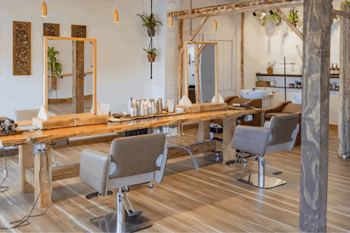 A bright, modern hair salon with wooden styling stations, large mirrors, and comfortable gray chairs. The space features natural wood accents, hanging plants, and organized shelves with hair products, creating a warm and inviting atmosphere.