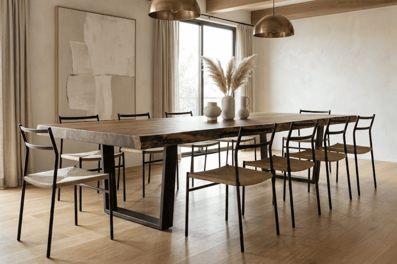 Sunlit minimalist dining room with a live-edge wood table, eight woven-seat chairs, pampas grass vases, pendant lights, and light curtains