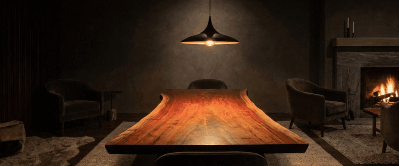 Warm, dimly lit dining room with a long live-edge wooden table, pendant light above, cozy armchairs and a crackling fireplace
