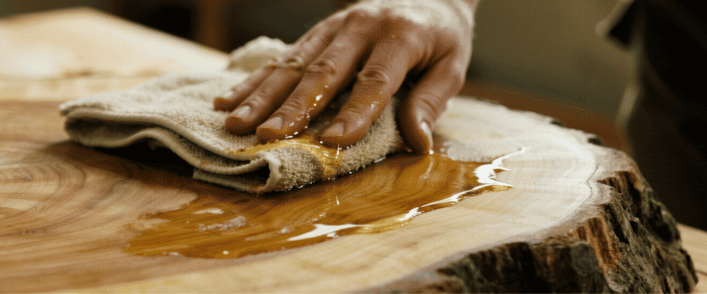 Close-up of a hand applying oil finish to a teak live-edge table with a lint-free cloth.