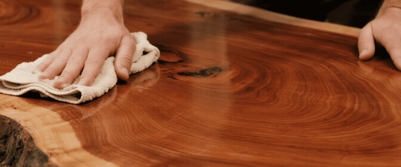 Man polishing a glossy live-edge wooden table with a cloth, showing hands, wood grain rings, and natural bark edge.